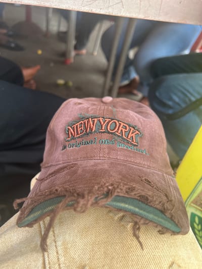 An overhead, angled shot captures a worn brown baseball cap embroidered with "NEW YORK" in two-tone lettering (dark green outline, orange fill), resting atop a person's lap. The cap's brim is frayed and tattered, revealing the green lining. The person's clothing includes what appears to be tan denim on the right side of the frame and a dark fabric (possibly black pants) beneath the cap. The background is indistinct, but there's evidence of what appears to be interior space, including metal frame chairs/seats and glimpses of other people's legs and feet. Based on the fact the hat is a New York themed hat, and the requested location is Aso Nigeria, there is not sufficient information to conclude where this picture was taken, only where it may be located based on the information provided.