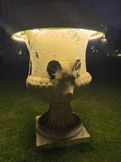 The image shows a large, weathered stone urn situated on a grassy area. The urn is brightly lit from within, with a light source visible around its upper rim. The surface of the urn appears textured and aged. The urn is likely located in Kathmandu Metropolitan City, Nepal. The time of day is night. The surrounding environment includes grass and blurred lights in the distance. There are no people visible in the image. The urn itself seems to be the main object of interest.