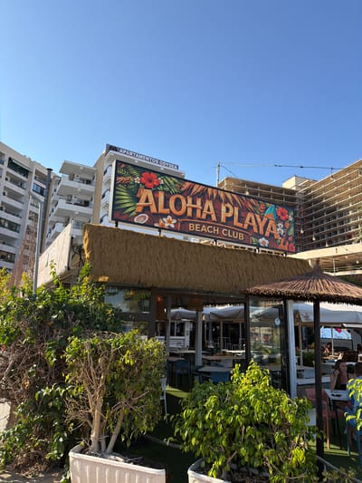 The image depicts a bright, sunny day in what appears to be a resort area. The scene is dominated by a large, colorful sign that reads "ALOHA PLAYA BEACH CLUB" in large, stylized lettering. The sign features tropical imagery, including hibiscus flowers, palm leaves, and a ukulele, suggesting a vacation or leisure setting.
In the background, there are tall, modern apartment buildings, one of which is under construction. The beach club itself has a thatched roof and outdoor seating areas visible through its windows. There are some people seated at tables outside the club, enjoying the weather. Green foliage from trees and bushes adds to the vibrant outdoor atmosphere.
The sky is a clear, vivid blue, indicating good weather and likely midday. The location is identified as Urbanització Cales I Talaies, Spain. The overall mood conveyed is relaxed and cheerful, typical of a beachside destination. There is also a smaller sign visible on one of the buildings that reads "APARTAMENTOS ODYSEA".
