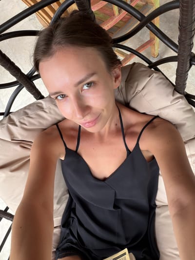 A woman with brown hair and green eyes is lying back in a wicker chair with a light brown cushion. She is wearing a black dress with thin straps. The chair is situated on what appears to be a patio or outdoor area, based on the presence of a painted wooden structure in the background. There is no information available to determine if the photo was taken in Shchelkovo, Russia.