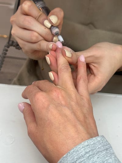 In Tunbridge Wells, United Kingdom, a close-up shot focuses on a person's hands as they undergo a manicure. One hand, held steady, displays nails painted in a pale pink and beige polish. The other hand, belonging to the manicurist, uses an electric nail file. The file is actively shaping or smoothing a nail on the client's hand, with fine dust particles visible in the air around the area of work. The background is blurred, suggesting an indoor setting, possibly a salon.