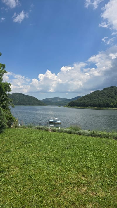 The image depicts a serene landscape scene, likely from Asparuhovo, Bulgaria, featuring a calm body of water bordered by verdant, tree-covered hills under a partly cloudy blue sky. In the foreground, a neatly manicured green lawn stretches towards the water's edge, which is lined with low-lying vegetation. A small, light-colored boat with a white canopy is anchored on the lake, appearing motionless. The overall atmosphere is tranquil and natural, suggesting a peaceful, outdoor setting.