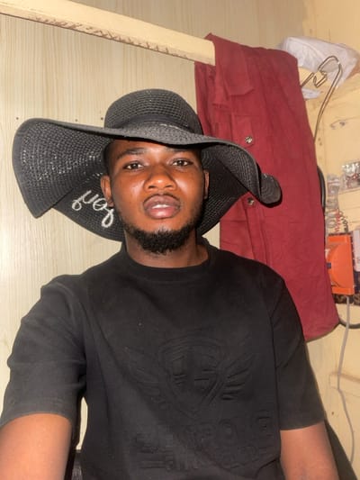 A man is lying on a wooden surface. He wears a black t-shirt with an embossed design and a black sun hat. The hat has a word embroidered on its brim. Behind him is a red garment or cloth piece, possibly on a bed. In the background, miscellaneous items are visible, including some white plastic bags and electrical wiring. The man appears to be posing, looking directly at the camera.