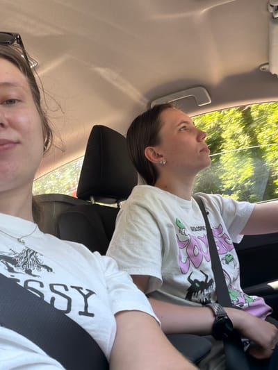 Two young females are seated inside a vehicle. The female in the foreground, on the left, is partially visible, with her face turned towards the camera. She is wearing a white t-shirt with a graphic print and a black seatbelt. Her sunglasses are resting on her head. The second female, on the right, is looking upwards and to the right, out the window. She is also wearing a white t-shirt with a graphic print and a black seatbelt. Her arm is resting on her lap, and she is wearing a watch. The interior of the car, including the headrest of a car seat, is visible. The exterior environment, consisting of green foliage and trees, is blurred due to the motion of the vehicle. The lighting suggests it is daytime, with sunlight filtering through the car windows. No specific location in Cannon Beach, United States is identifiable from the image.