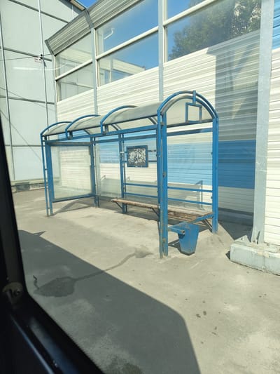 A bus stop shelter with a blue metal frame and a transparent roof is visible. The shelter has glass panels on its sides and front, with a wooden bench inside. A blue trash can is placed next to the bench. The ground is paved with asphalt, and there are some water stains and shadows. In the background, there is a building with a ribbed facade in white and blue stripes and large windows. A security camera is mounted on a pole on the left side of the frame. The scene appears to be a street or public area in the city of Zhukovsky, Russia.