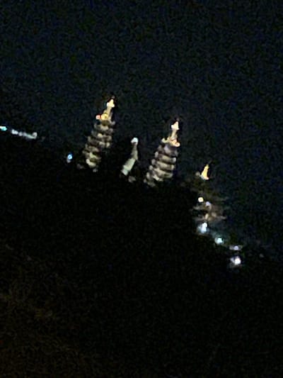 In the city of Phan Thiết, Vietnam, the image depicts a nighttime scene dominated by the dark silhouette of a hill against a black sky. On the slope of the hill, several illuminated pagoda-like structures are visible. The largest structure, positioned towards the left, is tall and segmented, with distinct layers of lighting creating a striped effect. To its right, a slightly smaller, similar structure also displays segmented lighting. Further to the right, another pagoda-like form with lighter, more diffuse illumination is present. In the foreground and scattered across the lower portion of the image, numerous small, bright lights suggest a landscape dotted with more lights, possibly from other buildings or streetlights. The overall impression is one of a nighttime cityscape viewed from a distance, highlighting illuminated architectural features on a natural elevation.