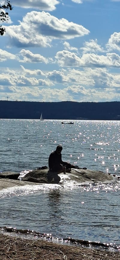 The image depicts a tranquil scene by a body of water, likely a lake or bay, under a sky filled with dramatic white clouds against a blue background. In the foreground, a person is seated on a rocky outcrop at the water's edge. The individual is silhouetted against the bright, shimmering surface of the water, which is reflecting the sunlight. The person appears to be wearing dark clothing and is looking down, possibly engaged in thought or holding an object. Beyond the seated figure, the water stretches out to a dark, forested shoreline in the distance. Several boats, including a sailboat and what appears to be a canoe or kayak with occupants, are visible on the water, adding a sense of scale and activity to the serene landscape. The overall impression is one of peaceful solitude and natural beauty. While the prompt mentions Lahti, Finland, there are no specific visual cues within the image that definitively confirm this location.