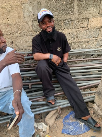 This image captures two adult males outdoors in Okene, Nigeria. The primary subject, positioned right of center, is a young adult male with a dark beard, wearing a white and black baseball cap, a dark ribbed polo shirt with "Reebok" branding, dark trousers, and black slides. He wears a dark digital smartwatch on his left wrist and has his left hand in his pocket. He is seated on a horizontal stack of metal pipes, smiling directly towards the camera. His skin exhibits some lighter discoloration on his forearms.
Partially visible on the left is a second adult male, facing towards the primary subject. He wears a white t-shirt and light blue denim jeans. His right hand is raised and open, gesturing as if in conversation. A light-colored bracelet with multiple small beads is on his right wrist, and he holds a light-colored smartphone in his left hand.
The background consists of a rough, light brown concrete block wall. The men are seated on what appears to be a haphazard stack of metal pipes or scaffolding tubes. The ground beneath them is dirt, littered with small rocks, pieces of broken concrete, crumpled white debris, and a flattened, crumpled blue plastic sheet.