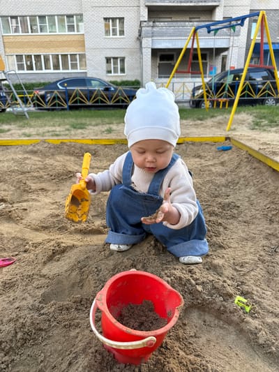 A baby wearing a white hat, a beige sweater, and blue overalls sits in a sandbox, playing with a yellow shovel and a red bucket filled with sand. The baby's eyes are closed, and their right hand is outstretched, holding the shovel. The background shows a playground structure and a building with windows. There are also cars parked near the building. The scene appears to be outdoors, likely during daytime, with natural light illuminating the sand. The ground is covered in sand, with some grass visible at the edges of the sandbox. The overall atmosphere is one of a child enjoying playtime in a playground. The image is oriented vertically, with the top of the frame showing more of the playground and building, and the bottom focusing on the baby and sandbox. There is no visible text in the image.