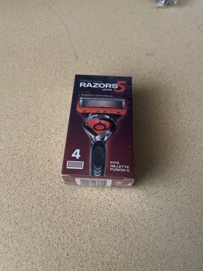 A top-down view shows a box of RAZORS 5 ULTRA razor cartridges on a speckled, beige carpeted surface. The box is dark red and black with a picture of a razor and lettering in Russian and English. Text on the box includes "Сменные кассеты" (Replacement cartridges), "RAZORS 5 ULTRA", "с лезвием-триммером" (with a trimmer blade), "4" with an image of a razor cartridge, and "FITS GILLETTE FUSION 5". The box appears to be sealed with plastic. The carpet has a coarse texture. The lighting suggests an indoor setting. There is no indication of the city of Oryol, Russia, in the image or its context.