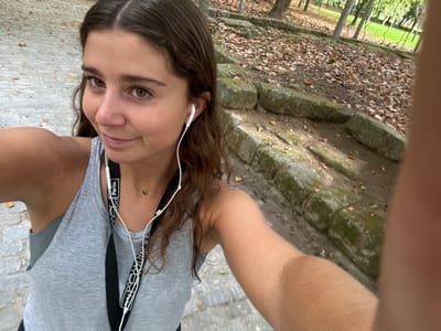 A young female, positioned centrally and angled slightly, captures a close-up selfie in an outdoor park in Aldoar, Foz do Douro e Nevogilde, Portugal. She has medium-brown wavy hair, dark eyes, and a subtle smile, looking directly at the camera. She wears a grey ribbed tank top over a darker undergarment, with white wired earphones inserted into both ears. A black lanyard featuring "Porto." text and a geometric pattern in white is visible around her neck, entwined with the earphone cable. A delicate gold-colored necklace with a small dark spherical pendant is also worn. Her right arm is extended, holding the camera, with portions of her left hand visible at the bottom right. The immediate foreground shows a grey cobblestone path. The background on the right reveals a dirt embankment featuring several layers of moss-covered, rough-hewn stone blocks. The ground of the embankment and portions of the path are covered with scattered dry brown leaves. Trees with green and sparse foliage are visible further back, beyond which a patch of green grass indicates an open area. The scene is illuminated by natural daylight.