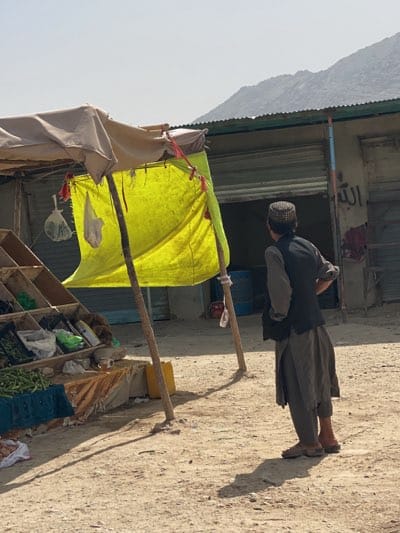 A young man wearing a black vest and a traditional cap stands near a roadside stall in Arghandab, Afghanistan. The stall is covered by a makeshift canopy with a yellow tarp. The stall displays produce. Behind the stall is a building with a metal shutter and Arabic script on the wall. In the background, a mountain is visible. The ground is dirt.