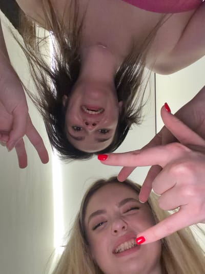 Two young women are angled down at the camera. One has dark hair, fair skin, and wears a pink top. The other has blond hair and fair skin. Both are smiling. Their hands are near their faces, posed with red painted nails. The background is blurred, showing a light color. The image has an amateur feel due to the angle and lighting.