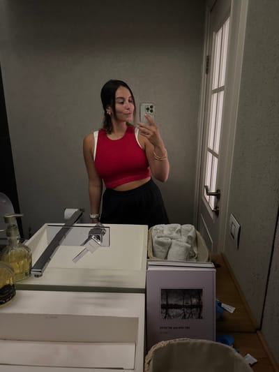 The image shows a young woman standing in front of a bathroom mirror. She is wearing a red tank top and black pants and holding a smartphone. She is looking at the camera. The bathroom is decorated in a modern style, with white walls and a white countertop. The countertop has a sink with a chrome faucet and a white soap dispenser. There is a wicker basket on the countertop with a towel inside. A book is sitting on the counter with a photo of a lake in the background. The woman is standing near the bathroom door. The image was taken in Aseret, Israel.