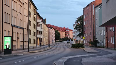 This is a photograph of a street scene in Linköping, Sweden, taken at dusk. The street is lined with multi-story apartment buildings on both sides. The buildings are of varying architectural styles and colors, including pale yellow, orange, and red brick. Many of the buildings have balconies. A street lamp is visible on the left side of the road, and another further down the street. On the left side of the image, there is a brightly lit digital advertisement board displaying a man in a green jacket and a white cap, with Swedish text. The road is asphalt, with white lane markings. Pedestrian crossings are indicated by white lines and triangular signs. There are trees and bushes visible along the sidewalks and between buildings. The sky is a pale blue, indicating the transition from day to night. The image captures a quiet urban street with a sense of calm and stillness.