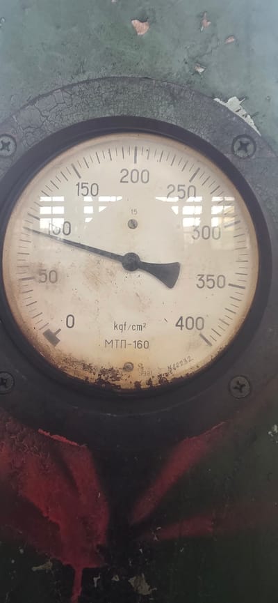 The image shows a close-up of a vintage pressure gauge on a weathered green surface. The gauge has a white face with black markings and numbers, indicating pressure in kgf/cm². The needle is pointing slightly above the 100 mark, indicating a pressure reading of approximately 110 kgf/cm². The gauge also has some text on it, including "kgf/cm²" and "МТП-160". The surrounding green surface is cracked and peeling, suggesting age and exposure to the elements. There are some red spray-painted markings on the lower part of the image, on a dark surface. The overall impression is of an old, industrial piece of equipment. There is no indication of the location being Karnobat, Bulgaria, or any specific city.