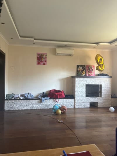 This is an indoor image of a living room. In the foreground, a wooden floor is visible. Towards the center of the image, a fireplace made of white stone is present with a black hearth. Above the fireplace, two framed pictures of animated characters are displayed. To the right of the fireplace, a large, gold balloon shaped like the number "3" is floating. On a ledge to the left of the fireplace, several items of clothing are laid out, including a red duffel bag. Further left, a painting of a tiger and strawberries hangs on the wall. In front of the ledge and fireplace, three decorative spheres are placed on the floor. A white, round object is also visible on the floor to the right of the fireplace. An air conditioner is mounted high on the wall above the paintings. The room has a modern ceiling with recessed lighting. The overall impression is that of a lived-in space, possibly decorated for a celebration given the balloon and artwork. The location is specified as Orekhovka, Russia, implying this is a residential interior in that city.