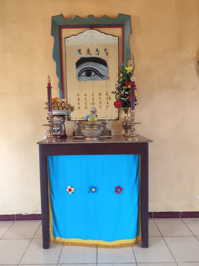 A table with a marble top is set up against a pale yellow wall. On the table, an altar is arranged with religious items. A framed painting of an eye with Chinese calligraphy is mounted on the wall above the altar. To the left of the painting is a red candle in a decorative holder, and a bowl of bananas. To the right of the painting, a vase holds a bouquet of colorful flowers. In front of the altar, the table is draped with a bright blue cloth adorned with three felt flowers: a yellow and red one on the left, a blue one in the middle, and a red one on the right. The cloth is trimmed with yellow fringe at the bottom. The floor is tiled with light-colored square tiles. The scene suggests a religious or spiritual space, possibly in the city of Lộc Quý, Vietnam, given the mention of the location.