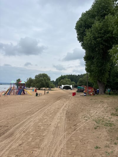 The image depicts a sandy area with tire tracks, suggesting vehicle access. In the background, a large, mature tree with lush green foliage dominates the left side of the frame. Beyond the tree, a white tent structure, possibly a stage, is visible. Several colorful children's playground structures are scattered across the sand, featuring slides and climbing frames. Some people are present, though not clearly identifiable, near the playground and a red structure. The sky is overcast with clouds. The location is identified as Zhdanovichi Village Council, Belarus. The scene appears to be a recreational area, likely a beach or park, with provisions for events and children's activities. The time of day is difficult to ascertain due to the overcast weather, but it appears to be daytime. There is no visible text in the image.