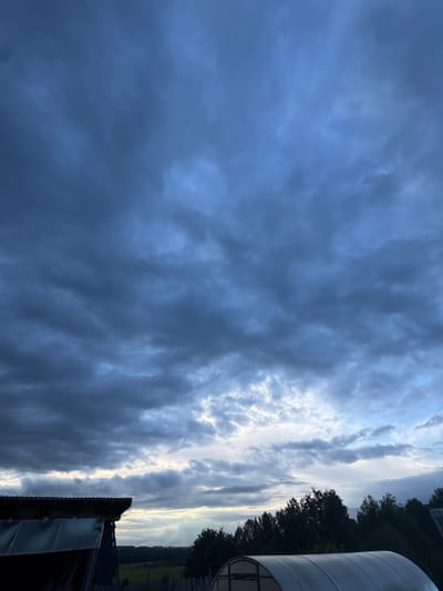 The image shows a cloudy sky over Nizhnyaya Gar', Russia. The sky is predominantly filled with dark, grey clouds on the left side. A bright, narrow opening of lighter, white clouds cuts through the center of the image. The right edge reveals dark green trees and a section of a white, cylindrical structure. A small portion of a roofline is visible in the lower right corner.