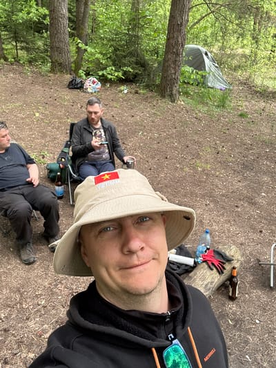 The image shows three individuals in a wooded area. A man in a black hoodie and a tan hat with a Vietnamese flag patch is taking a selfie. Behind him, a man is sitting in a camping chair, looking at a smartphone. Another man is lying on the ground on a small camping chair. A tent is visible in the background near the left side of the image. Other camping paraphernalia such as bottles, gloves, and a small stove can be seen scattered in the area. The ground is covered with dirt and dead leaves. The location is Юзуфоўскі сельскі Савет, Belarus, based on the description.