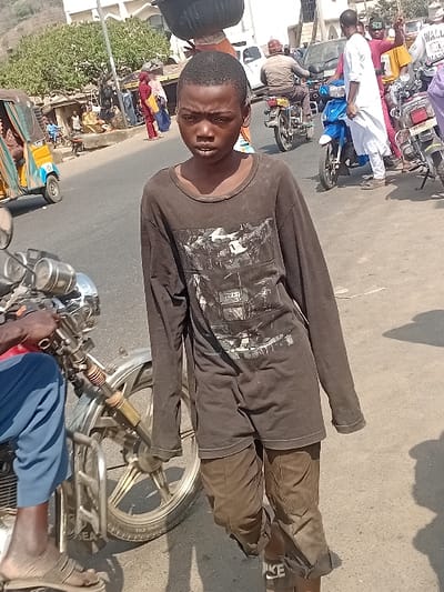 A young man in a dark long-sleeved shirt and pants is standing in front of a parked motorcycle. He appears to be on a street, with other motorcycles and people visible in the background. The scene is outdoors on a sunny day. He is wearing sandals and looking at the camera. Buildings and vehicles are seen blurred in the distance.