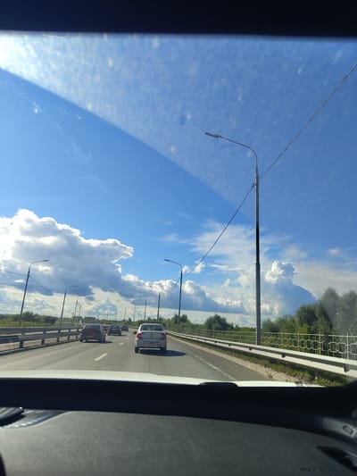 The image captures a view from inside a vehicle looking out through the windshield at a highway scene in Фруктовая, Russia. The sky is predominantly blue with large, puffy white clouds. Several streetlights line the right side of the highway, and a power line is visible running diagonally across the sky. Trees and foliage form a green border along the right side of the road. Multiple cars are driving in the same direction on the highway, with traffic appearing moderate. The dashboard of the vehicle is visible at the bottom of the frame. The overall impression is a sunny day on a suburban or rural highway.