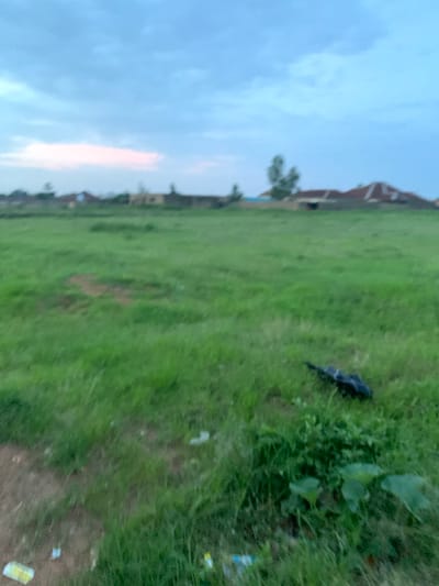 The image shows an outdoor scene with a wide expanse of green grass under a cloudy sky. In the background, there are several buildings with reddish-brown roofs, suggesting a residential area. A few trees are also visible. In the foreground, there is a black plastic bag lying on the grass. The scene appears to be in Du, Nigeria, judging by the context provided. The overall atmosphere is one of an overcast day in a somewhat rural or semi-urban environment.