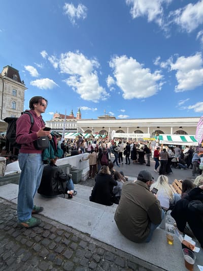 The image depicts a sunny, outdoor scene in Ljubljana, Slovenia, likely taken in or near the city's central marketplace. In the foreground, a young man stands wearing a maroon sweatshirt, jeans, sunglasses, and green sneakers. He carries a black bag and holds a device, possibly a phone, in his hand.
Behind him, a crowd of people is gathered along a building with a long, white arcade structure. There are food stalls under white awnings. People are sitting on the steps and ledges. The marketplace seems to be crowded with people.
Above the buildings, a bright blue sky is visible with scattered white clouds. The buildings feature architectural details typical of Ljubljana, including turrets and ornate facades. The ground is paved with cobblestones.