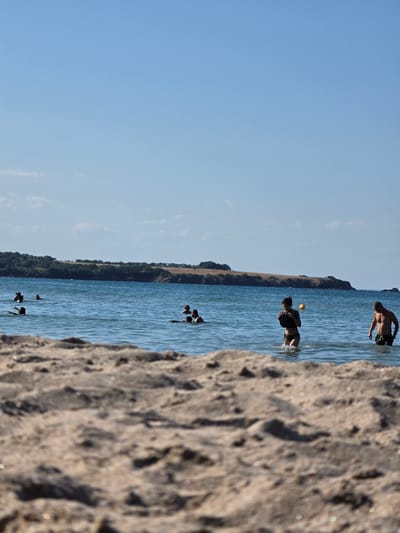 The image depicts a beach scene in Ahtopol, Bulgaria. In the foreground, the sandy beach is blurred and out of focus. The midground shows a body of water with several individuals swimming and wading. On the right side of the water, a man in dark swim trunks stands partially submerged, facing left. To his left, a woman in a black bikini is wading out of the water, holding a black object to her chest, with a yellow ball visible in the water to her right. Further out in the water, other people are visible, some swimming and one person on a surfboard. In the background, a long, treed coastline stretches across the horizon under a clear blue sky with a few scattered clouds. The sea is calm with gentle waves.
