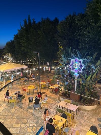This image captures an outdoor seating area at night, likely a restaurant or cafe, with numerous tables and chairs. The setting appears to be in Barletta, Italy, as indicated by the presence of a decorative flower-shaped light installation with purple and blue LEDs, and a white pole structure.
In the foreground, several people are seated at tables, engaged in conversation and dining. One woman in the lower right is standing and appears to be talking to a man. There are also individuals seated at tables in the midground and background.
The environment is illuminated by various light sources, including string lights overhead, table lamps, and the prominent decorative light fixture. Large trees with dark foliage form a backdrop, suggesting an outdoor courtyard or garden space. The ground is paved with stone tiles. The overall ambiance is lively and social, typical of an evening gathering at an establishment.