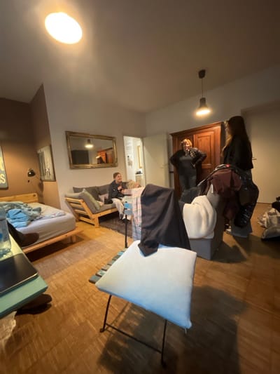 The image captures an indoor residential space, identified as being in Prague, Czechia. The room features light-colored wooden flooring, white walls, and a dark brown-grey accent wall section on the left. Two overhead lights provide illumination: an oval fixture on the left and a pendant light with a black shade on the right.

Four individuals are present, engaged in a casual social interaction. In the mid-ground left, a young female with dark hair, wearing a dark hoodie, is seated on a light wooden sofa with grey cushions, smiling and holding a small object. Another individual with light hair is partially visible beside her on the sofa. In the central-right area, two females stand facing each other. One, with reddish-brown hair, wears a dark jacket and dark trousers, standing with her hands on her hips. The other, with long dark hair, wears a dark jacket and dark trousers, looking towards the first woman.

The room's furnishings include a wooden bed frame with light blue bedding and blue clothing draped on it, located in the foreground-left. A small, light blue-green table with a plastic water bottle and a dark electronic device is positioned in the immediate foreground-left. A metal chair with a light blue cushion and a black garment draped over its back stands in the mid-ground center. To its right, a grey armchair is covered with various clothes, including a white knit sweater, a black puffy jacket, and a brown garment. A large, ornate gold-framed mirror hangs on the back wall, reflecting part of the room. Adjacent to the mirror, a dark wooden double-door wardrobe stands prominently. A doorway leading to an adjacent room is visible to the left of the wardrobe. Additional clothing items are piled on the floor in the far right of the image.