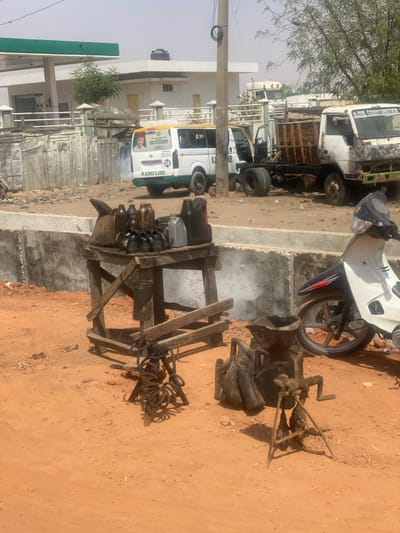 The image captures an outdoor roadside scene in Bachirawa, Nigeria, under bright, clear daylight, showing reddish-brown dirt ground throughout.
In the foreground, a crudely constructed wooden table stands, laden with an assortment of containers including dark plastic bottles, metal cans, and various small, dark mechanical components. Beneath the table, a dark bucket is suspended, and on the ground to the left, a cluster of metal coils or springs is present. To the right of the table, two weathered, dark metal objects rest on the dirt: one is an accumulation of coiled metal springs, and the other is a larger device with a funnel-shaped top and a hand-crank mechanism, possibly a manual oil or fluid pump. The front section of a white and black scooter with a brown wheel is partially visible on the far right edge of the frame.
A low, concrete wall serves as a barrier, separating the immediate foreground from the mid-ground. Beyond this wall, two vehicles are parked on a similar dirt surface. On the left, a white passenger van with green and yellow stripes bears "KANO LINE" text and a political campaign poster featuring a man's portrait on its rear window. To its right, a white flatbed truck appears partially disassembled, with its passenger-side door missing and a wooden cargo frame on its bed. A dark utility pole is positioned vertically between these two vehicles.
The background consists of a white building with a prominent green-canopied structure, indicative of a gas station. A black water tank is visible on the roof of the white building. Sparse, light-colored trees and dry vegetation are also visible in the background. No individuals are present in the scene.