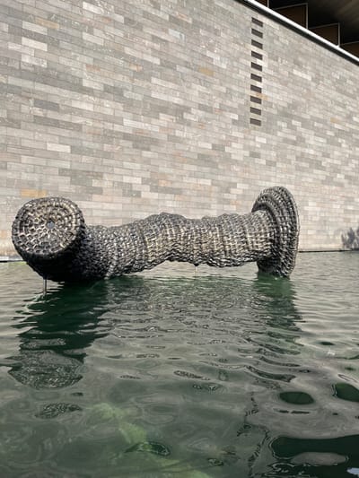 The image captures an outdoor scene dominated by a large, dark-colored sculpture positioned between a paved walkway and a body of water, bathed in bright daylight.
The main subject is an elongated, stylized sculpture resembling a giant, intricately woven handle or a basket-like form. It features a cylindrical shape, with wider, flatter ends and a wavy, undulating mid-section. The sculpture's surface exhibits a dark, metallic sheen with visible woven textures, suggesting it is crafted from interwoven strips of material, possibly metal or a durable synthetic, mimicking wicker. Small, slender metallic supports are visible where the sculpture meets the water, indicating it is an art installation.
To the left, a spacious paved area stretches out, likely a public plaza or walkway. It is surfaced with numerous light grey rectangular pavers, arranged in a staggered, brick-like pattern, with slight variations in tone and texture. Along the top-left edge of this paved expanse, a glimpse of a building or structure is visible, featuring a series of dark, rectangular grilles or vents embedded within its base.
To the right of the sculpture, a calm body of water extends across the frame. Its surface, dark green-grey in hue, displays gentle ripples, reflecting subtle patterns of light and shadow, possibly from the sky or surrounding foliage. The sculpture casts a prominent, elongated shadow onto the water, underscoring the bright and clear sunlight.
The scene is devoid of any people, creating a serene and quiet atmosphere. The strong, crisp shadows and overall illumination suggest it is mid-morning or early afternoon on a sunny day with clear weather. The setting appears to be a modern urban or public space, consistent with the context of Southbank, Australia, likely a riverfront or waterside development featuring public art. There is no visible text in the image.
