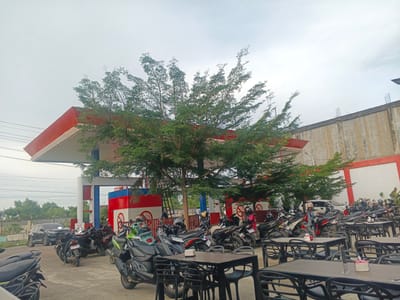 The image depicts a gas station with numerous motorcycles parked in front of it. The gas station has a distinctive red and white canopy and red and blue striped pillars. Several trees with lush green foliage are planted around the gas station, providing shade. In the foreground, there are tables and chairs, suggesting an area for customers to rest or dine. Several motorcycles are parked in rows, with some having helmets on them. A black car is parked on the left side of the frame. The background shows a building with a concrete wall and some electrical wires.
The scene appears to be set during the daytime, with an overcast sky, indicating possibly cloudy weather. The presence of many parked motorcycles suggests that it might be a busy time at the gas station. The location is identified as Geulumpangsulu Timu, Indonesia.
There is some visible text on the gas station pumps which is difficult to read clearly due to the resolution, but one sign on a pump appears to say "PERTAMAX". There are also some circular signs with what looks like a "no smoking" symbol on them.