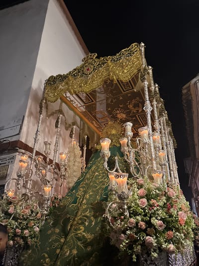The image depicts a religious procession float in Córdoba, Spain. The float features an ornate gold-trimmed canopy with a green velvet skirt adorned with gold embroidery. Silver candelabras with lit candles are visible. Bouquets of pink and white roses and other flowers decorate the float. A building is partially visible on the left side, with darkness above.