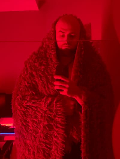 The image shows a person wrapped in a fluffy, red blanket, holding a mobile phone. The scene is lit with red light, creating a dim and moody atmosphere. The person appears to be male, with a beard and a shaved head. The background is out of focus, but suggests an indoor setting with some electronic equipment visible on the left side of the image. There is a faint horizontal line across the image, possibly from a reflection. The location of this image in Gliwice, Poland, cannot be determined from the visual information provided.