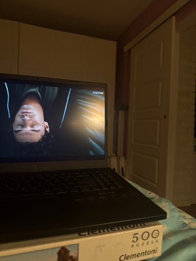 A person is lying on their back, with their head towards the left side of the frame, facing a laptop screen. The laptop is open and displaying a video of the person's face, viewed upside down, with their eyes closed. The laptop has a black keyboard and is resting on top of a puzzle box. The puzzle box is labeled "Clementoni 500 PUZZLE". The background of the image shows a closet door and a wall with white brick-like texture. The scene appears to be indoors, in a bedroom or living area. There is no indication of the city of Pokrovnik, Bulgaria in the image.