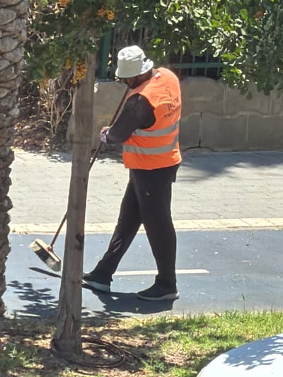 A person wearing a white bucket hat and an orange high-visibility vest with reflective stripes is sweeping the ground with a broom. They are wearing dark pants and dark shoes. The person is positioned near a thin, light-colored tree trunk, with a palm tree trunk visible on the left. The background shows a paved surface, possibly a sidewalk or bike path, with a low stone wall and green foliage beyond. The scene appears to be outdoors, in a public area. The person is right-handed and is sweeping from right to left. The area is well-lit by sunlight, casting shadows on the ground. The overall environment suggests a street or park setting.
