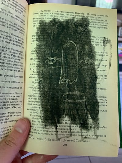 This is a close-up, high-angle shot of an open book with a drawing on the page. The drawing is a charcoal sketch of a face, rendered in a cubist style with prominent eyes and abstract lines. The sketch is smudged and covers a significant portion of the text, making some of it illegible.

The book appears to be an older edition, with yellowed pages. The text is in Russian and is printed in a serif font. The drawing has been applied directly onto the pages, obscuring the printed words and creating a layered effect.

The scene is dimly lit, suggesting an indoor setting, possibly a room in a house or a library. The background is out of focus, but hints of green and brown suggest a natural or organic environment. A person's hand, with a visible thumb and index finger, is visible at the bottom right corner of the image, holding the book open. The hand appears to be fair-skinned and is positioned as if the person is reading or examining the page.

The overall impression is one of artistic expression layered over literature, with the charcoal drawing dominating the visual space and transforming the printed text into a canvas. The smudged nature of the drawing suggests it was done with a soft medium like charcoal or chalk. The emotions conveyed are somewhat ambiguous due to the abstract nature of the drawing and the obscured text, but the intensity of the charcoal strokes could imply strong emotion or a bold artistic statement.