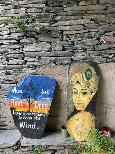 Two painted stones are displayed against a stacked stone wall in Bhagsu, India. The stone on the left is heart-shaped and painted with a sunset scene featuring a tree and the quote, "When the Root is Deep there is no reason to Fear the Wind." The stone on the right is rectangular and depicts a painted portrait of a person with a turban adorned with a peacock feather, holding a flute. Two empty tin cans and a small green plant are situated at the base of the stones.