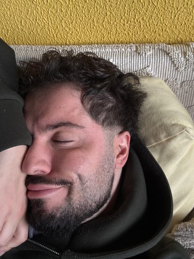 The image depicts a close-up, overhead view of an adult male lying on a textured surface, presumably a bed or sofa, in Vila Nova de Gaia, Portugal. The male has dark, wavy hair, a trimmed beard, and closed eyes, suggesting he is asleep or resting. A faint smile is visible on his face. His left cheek and nose area are partially obscured by a light-skinned hand, which appears to be pressed gently against him. He is wearing a dark, possibly black, hooded garment. Directly behind his head is a light-colored, textured pillow or cushion. Beyond that, a white or cream-colored textured fabric is visible, topped by a bright yellow, highly textured surface.