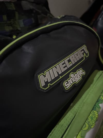 A close-up shot focuses on a black backpack, with a Minecraft logo and a Smiggle logo prominently displayed. The backpack has a green trim. A green pencil case rests on top of the backpack. The image was taken in Binong, Indonesia.