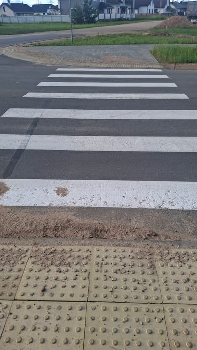 The image depicts a pedestrian crossing at street level in Stowbtsy, Belarus. The crossing is marked with white stripes on asphalt. In the foreground, tactile paving tiles are visible, designed to assist visually impaired pedestrians. These tiles are yellow and feature a pattern of raised domes. Sand and debris are present on the edge of the road and on the tactile tiles. In the background, a residential area with houses and green grass can be seen. A dirt pile is also present in the background. The sky is overcast. No people are visible in the image.