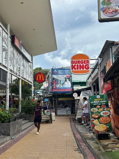 This image captures a street scene in Ao Nang, Thailand, featuring a row of businesses under a cloudy sky. On the left, a McDonald's restaurant is visible with its iconic golden arches sign. Further down the street, a prominent Burger King sign is displayed. To the right, a large green advertisement for "The Pizza Company" showcases images of pizza, pasta, steak, and salad. Several people are walking along the pedestrian walkway, which is paved with yellow and brown tiles and has a textured tactile paving strip. One man in a maroon t-shirt and black shorts is walking away from the camera, while another man in a white t-shirt and purple shorts walks towards the camera. The buildings lining the street appear to be a mix of commercial establishments with visible signage and awnings. Lush green foliage is present in some areas, particularly near the McDonald's. The overall atmosphere suggests a bustling commercial area, possibly a tourist destination.
