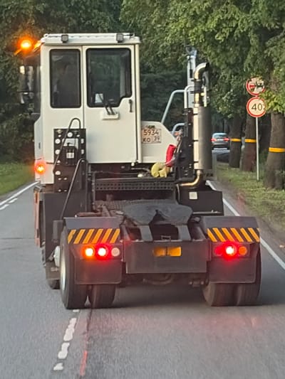 An outdoor, low-angle, eye-level, medium shot shows the rear of a white and black terminal tractor on a paved road. The tractor has a large white cab with two windows and an orange turn signal on the top left. The rear of the tractor features a dark gray fifth wheel and two sets of red brake lights with yellow and black striped reflective tape above them. A white license plate with Russian characters and numbers "5934 XO 39" is visible on the side of the tractor. On the right side of the road, there are trees with yellow bands around their trunks and two circular traffic signs, one depicting a red circle with a horizontal bar and a red diagonal bar, and another with the number "40" in a red circle. A silver sedan is visible in the distance on the road ahead. The scene appears to be in the city of Primorsk, Russia, likely during the day given the lighting.