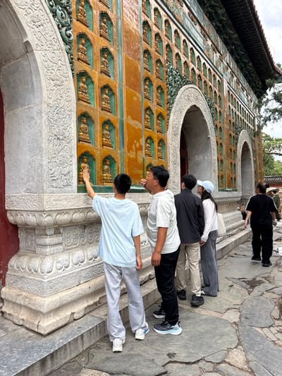 The image depicts an outdoor scene at the Hall of Ten Thousand Buddhas (Wanshou Mountain) within the Summer Palace (Yiheyuan) in Haidian District, Beijing, China.
A large, ornate building facade dominates the scene, characterized by its extensive wall decorated with a grid of recessed niches. Each niche, framed in green glaze and set against an orange-glazed tile background, houses a small, golden-colored, seated Buddha figurine. Three large, grey stone arched doorways are visible along the wall; their surrounds and the building's base are intricately carved with floral and scrollwork patterns. Decorative green elements are present above the arches and along the upper section of the wall, beneath a dark-tiled roof with extended eaves visible at the top right.
In the foreground, a young male, appearing to be between 8 and 12 years old, wearing a light blue t-shirt and grey sweatpants, stands with his back partially to the viewer, reaching his right hand to touch a Buddha figurine in a lower niche. To his immediate right, an adult male in a white t-shirt and dark pants observes the wall, looking upwards with his mouth slightly open. Further back, a male in a black jacket and tan pants stands next to a female wearing a white long-sleeved shirt, blue jeans, and a light blue baseball cap, both also looking up at the building. In the background on the right, an adult female in a black tracksuit walks away from the viewer along a stone path.
The ground is paved with irregularly shaped grey stone slabs, displaying cracks and uneven surfaces. Trees are visible in the background, and the sky appears overcast.