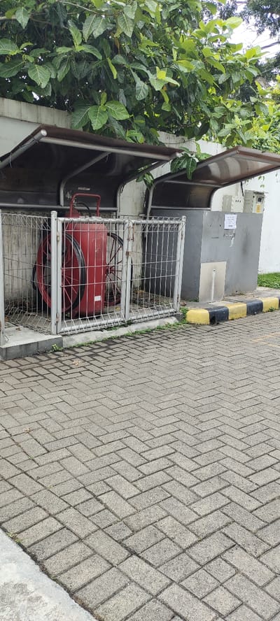 A red fire extinguisher is enclosed in a metal cage with a metal roof. The cage is situated on a brick walkway, surrounded by green foliage. The fire extinguisher is mounted on wheels and is not in use. The scene is located in Kecamatan Lawang, Indonesia, at latitude -7.85 and longitude 112.695. The environment appears to be a public area, possibly a school or business. There are no people present.