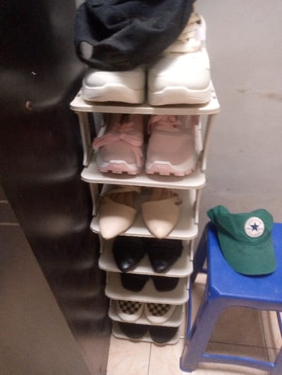 The scene depicts an organized indoor space, likely a residence or entryway in Cimenyan, Indonesia, though the exact location within the city cannot be determined from the image. A seven-tiered, white, modular shoe rack stands vertically against a light-colored wall, storing multiple pairs of footwear.
From top to bottom, the rack holds:
- The topmost shelf displays one pair of white athletic shoes, with a black fabric cap resting horizontally on top of them.
- The second shelf contains one pair of pink athletic shoes with white soles.
- The third shelf holds one pair of light beige, pointed-toe, low-heeled or flat shoes.
- The fourth shelf features one pair of black, pointed-toe flats or low-heeled shoes.
- The fifth shelf displays one pair of plain black slip-on shoes, possibly loafers.
- The sixth shelf contains one pair of white and black checkered slip-on shoes.
- The bottom shelf holds one partially visible pair of dark-colored shoes.
To the immediate right of the shoe rack, a short, four-legged blue plastic stool is positioned on the tiled floor. A green visor cap, featuring a white circular emblem with a blue star, rests on the seat of the blue stool. The flooring is composed of light-colored, possibly beige, square tiles with visible grout lines. A dark, vertical surface, possibly a door frame or furniture, is visible on the left side of the frame, partially obscuring the leftmost portion of the shoe rack. The background wall behind the rack and stool appears to be a light, solid color, exhibiting some minor discoloration or staining towards the right. No individuals are present in the scene, and no active interactions are occurring. The objects are static.