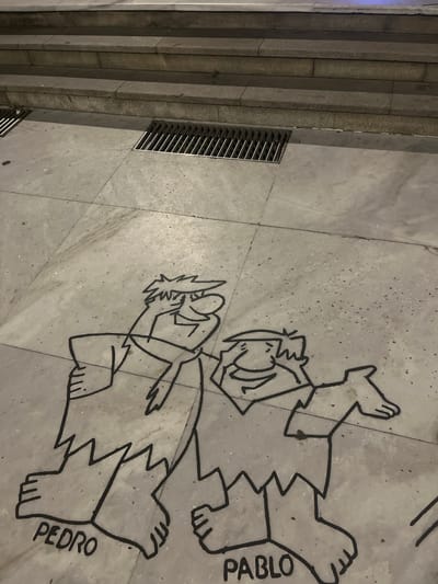 The image shows a street art depiction of characters from The Flintstones, specifically Pedro and Pablo, drawn in black on a concrete pavement. The characters are depicted in a simplified, outline style, with jagged edges on their clothing and feet. The word "PEDRO" is visible below the left character, and "PABLO" is visible below the right character. The pavement is segmented by grout lines, and in the background, there are stone steps leading upwards and a metal grate for drainage. This street art is located on Calle de la Marineda in A Coruña, Spain, near the Domus museum. The scene is outdoors, and the lighting suggests it is nighttime or a heavily shaded area.